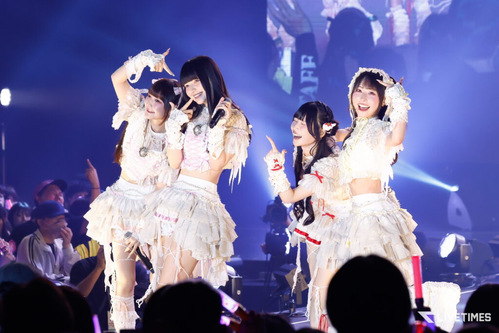 Four female performers in white frilly costumes pose and smile on stage during a live concert, crowd in the foreground.