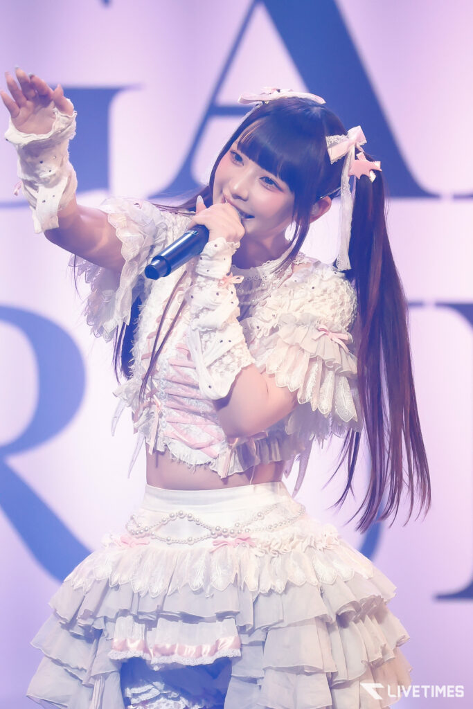 Young female singer in a frilly white-pink stage outfit holding a microphone and performing on stage.