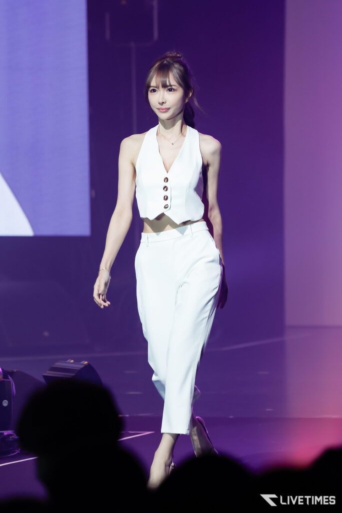 Woman in a white sleeveless top and white pants walking on stage under purple lighting, audience silhouettes in the foreground.