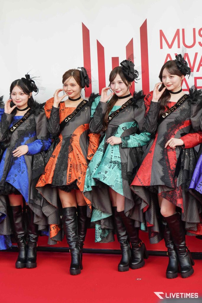 Four women in colorful, patterned dresses with black accessories and boots pose on a red carpet at a music awards event under white backdrop.