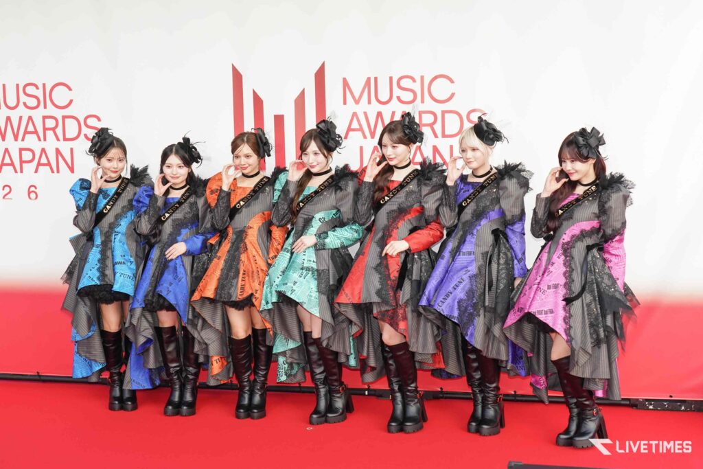 Nine women in colorful patterned outfits posing on a red carpet at a music awards event, with black boots and matching hats, LIVETIMES watermark bottom right