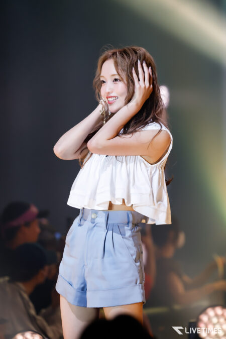 Performing woman on stage, smiling and touching her hair, wearing a white top and light blue shorts with a blurred audience behind her, LivesTimes watermark visible in corner.