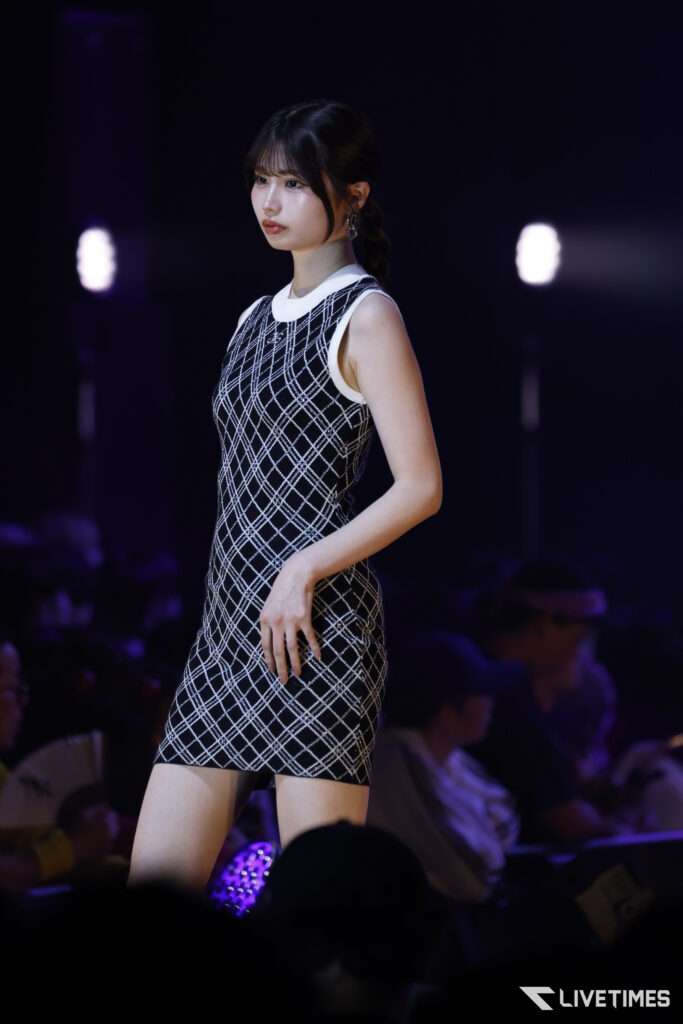 Female model walking a catwalk in a sleeveless black-and-white plaid dress under purple stage lights, with a blurred audience in the background.