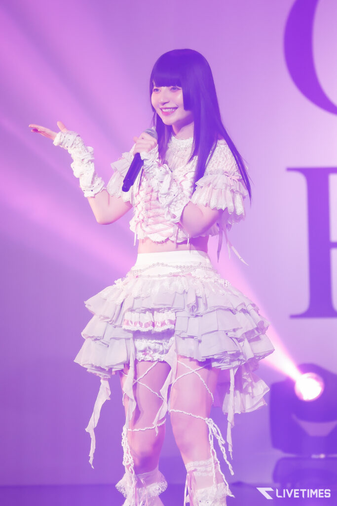 Female singer on stage in a frilly white outfit, holding a microphone and smiling under purple lights.