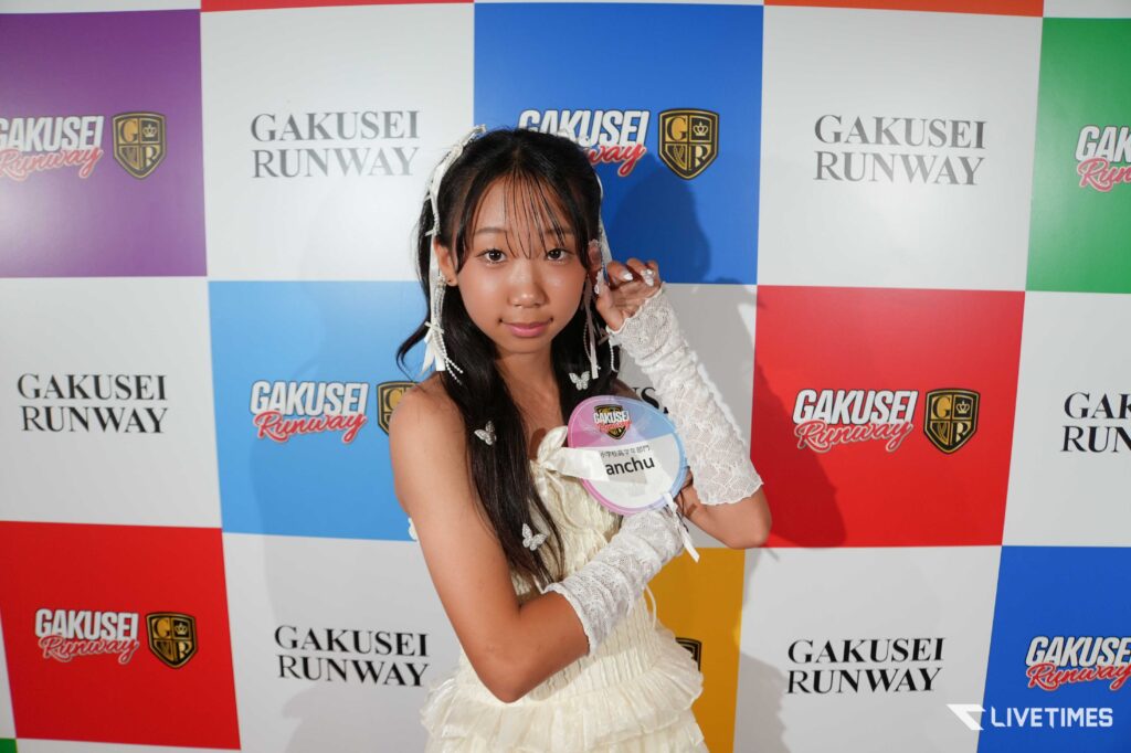 Young girl in a white dress posing at a colorful Gakusei Runway backdrop, holding a round badge.