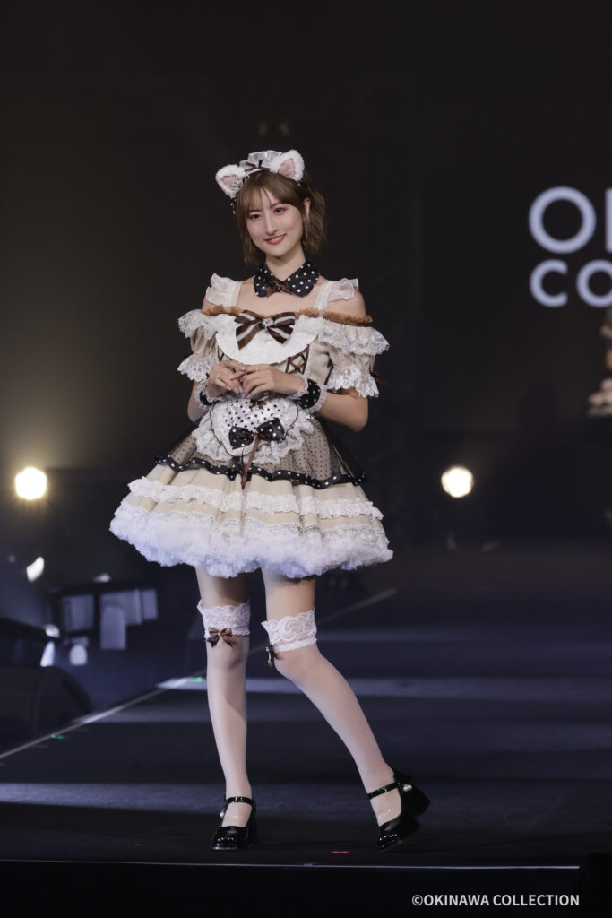 Woman on a dark stage in a frilly white-and-brown maid costume with lace, bows, and cat-ear headband, smiling at the audience.