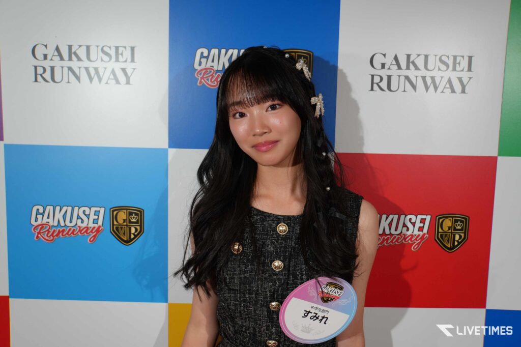 Portrait of a young woman with long dark hair in a black dress with gold buttons, posing in front of a colorful 'GAKUSEI RUNWAY' backdrop.