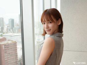 Woman with short brown hair standing by a large window, city skyline visible outside in the background.