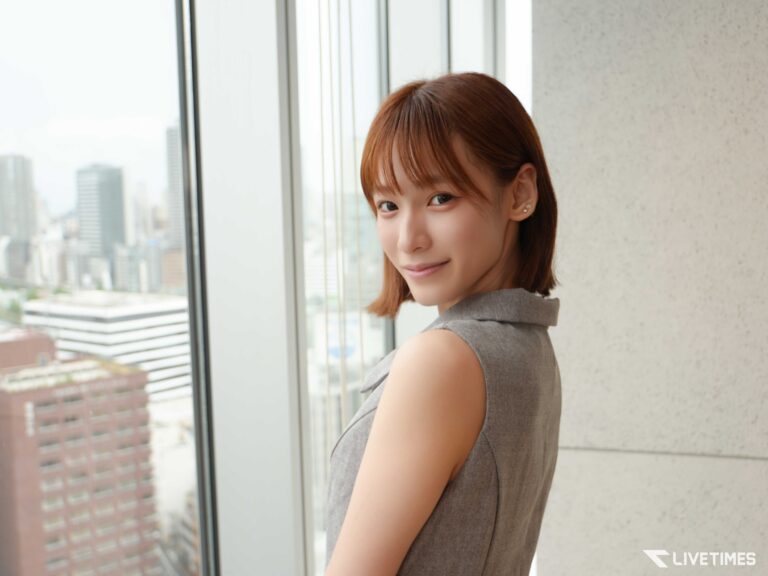 Woman with short brown hair standing by a large window, city skyline visible outside in the background.