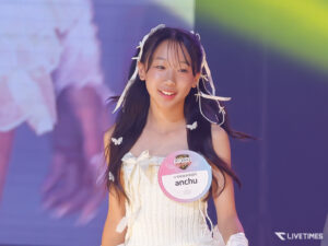 Young girl in a white dress on stage, smiling, with butterfly hair accessories and a pink badge reading 'anchu'.