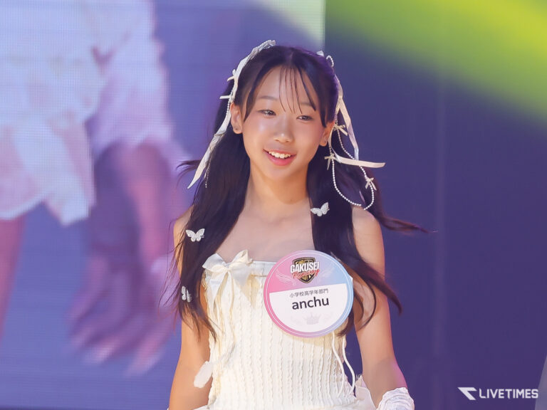 Young girl in a white dress on stage, smiling, with butterfly hair accessories and a pink badge reading 'anchu'.