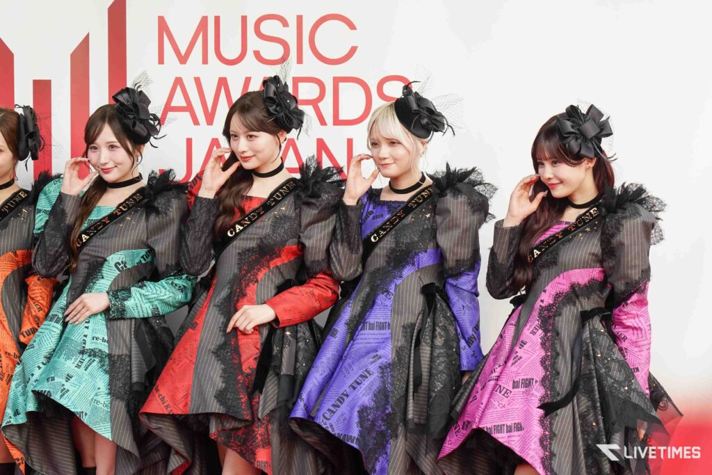 Five young women in coordinated, colorful dresses with black headpieces posing at a Music Awards Japan event.