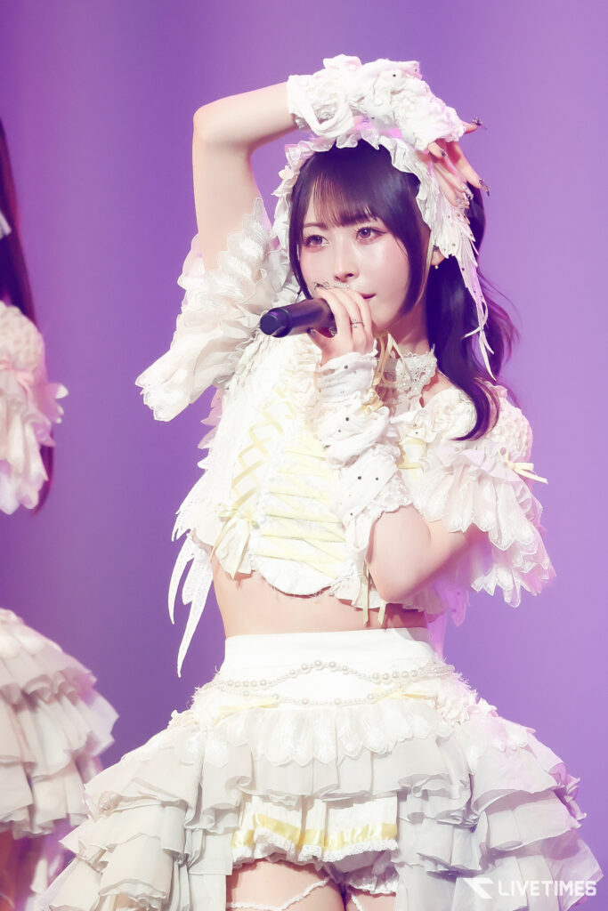 Singer performing on stage in a white, frilly dress with ruffles, holding a microphone under purple lighting.