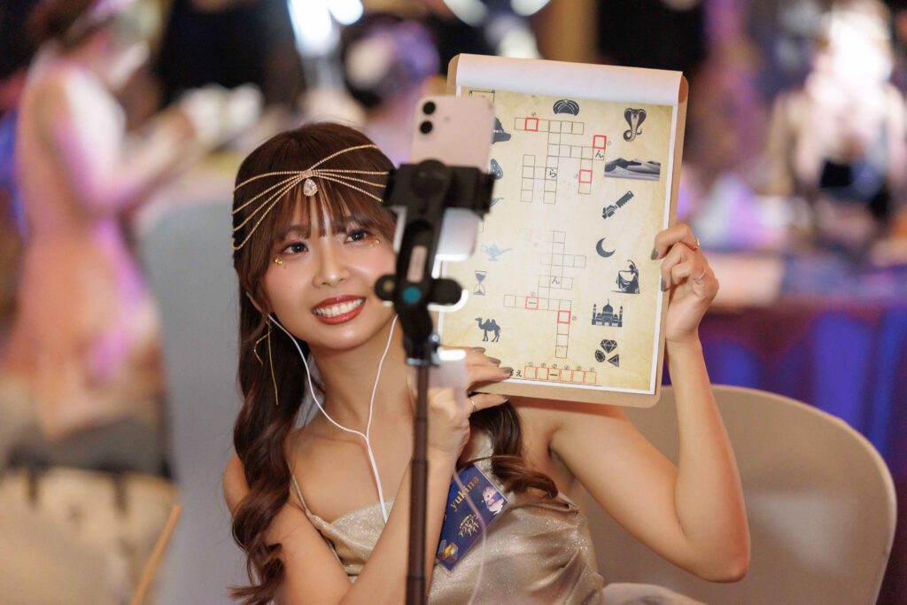 Woman with a jeweled headpiece smiles at a phone on a tripod while holding a decorated puzzle sheet nearby.