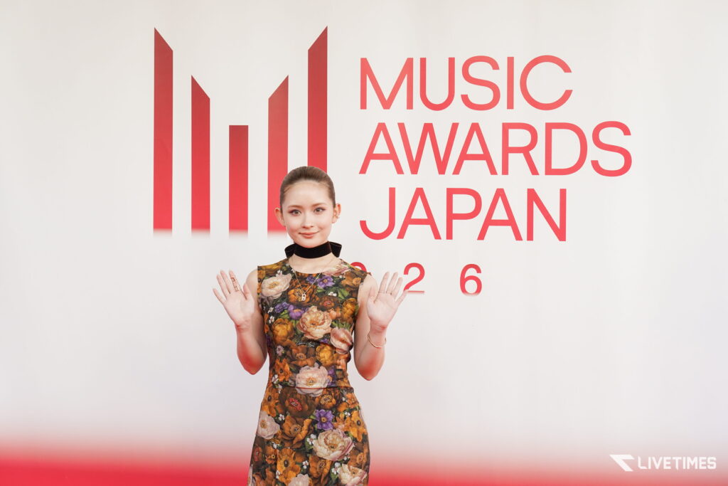 Woman in a floral dress poses with hands up in front of a large 'MUSIC AWARDS JAPAN' backdrop with red vertical bars on a white wall.