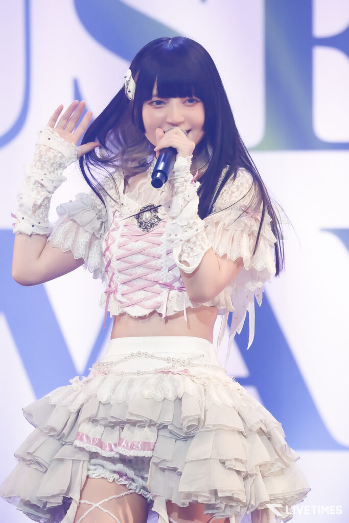 Young female singer in a frilly white lace stage outfit, waving while holding a microphone at a performance.