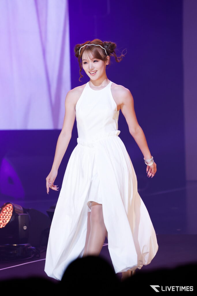 Female performer in a white sleeveless gown smiling on stage with purple lighting in the background.