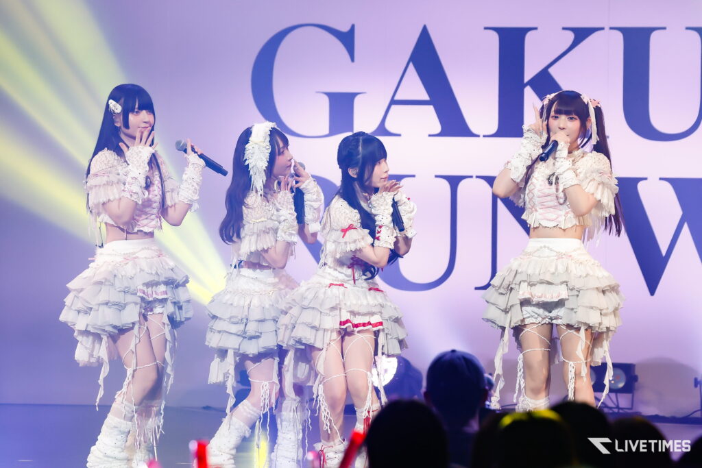 Five female pop artists in white frilly outfits perform on a stage with microphones, under colorful lights and a large backdrop behind them.