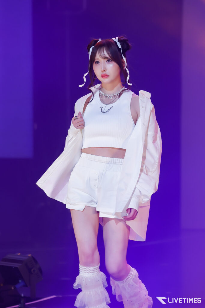 Stage performer in a white crop top and shorts, white jacket, with ribboned pigtails under purple lighting, center-stage.
