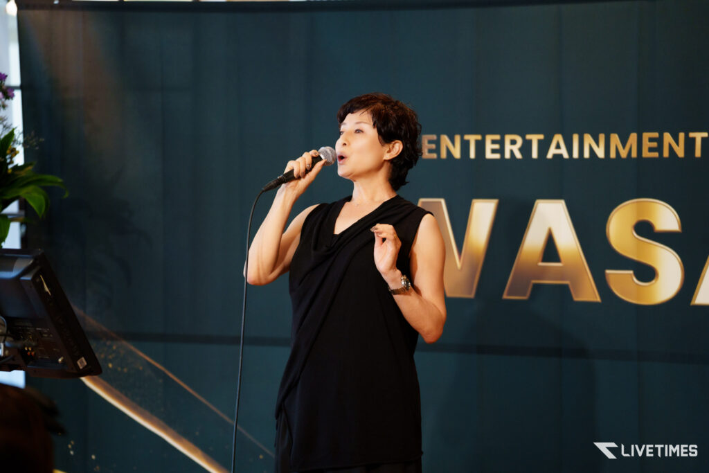 Woman singer performing on stage with microphone, dark outfit, against a backdrop that reads 'ENTERTAINMENT' and 'LIVETIMES' logo in the corner