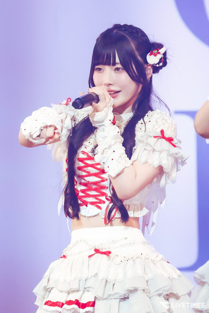 Young female singer performing on stage, holding a microphone in a white lace costume with red ribbons and hair ornament