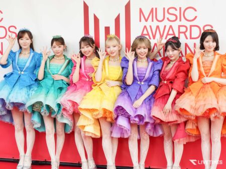 Eight women in rainbow-colored dresses with tiaras pose together on a red stage backdrop at a music awards event.