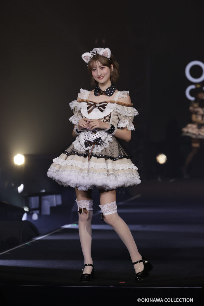 Stage performer in a cream lace dress with bows and a cat-ear headband on a dark stage.