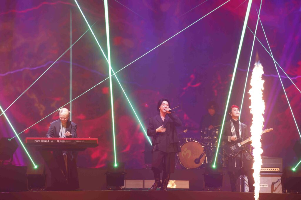 Live band on stage with neon lines, singer center, keyboardist left, guitarist right, flames rising nearby.
