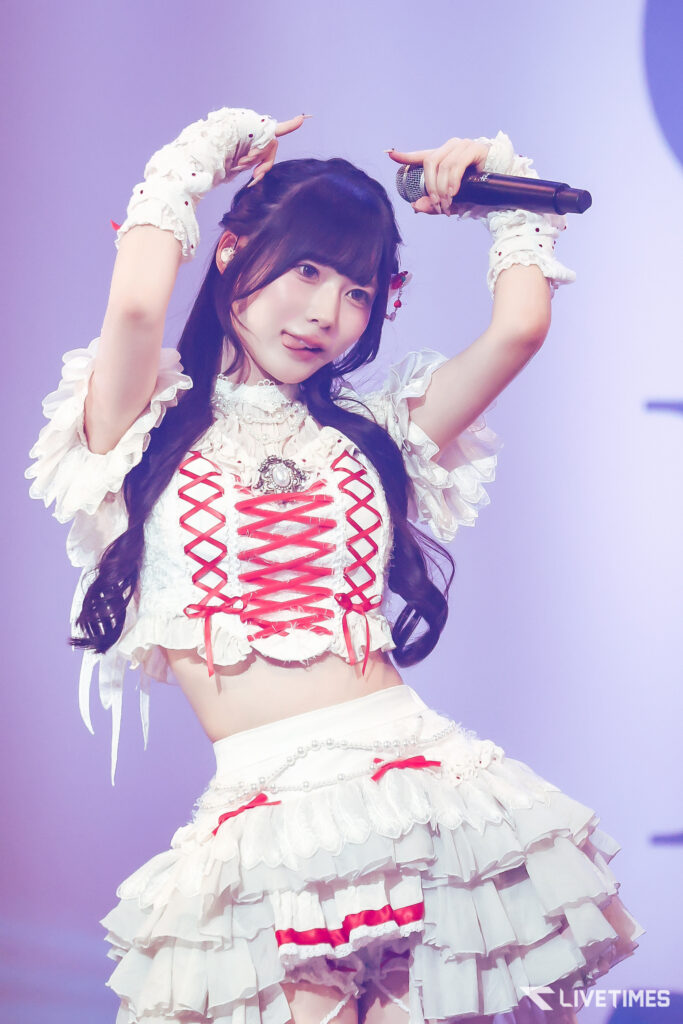 Female performer in a white frilly stage outfit raises her arms with a microphone against a purple background on stage.