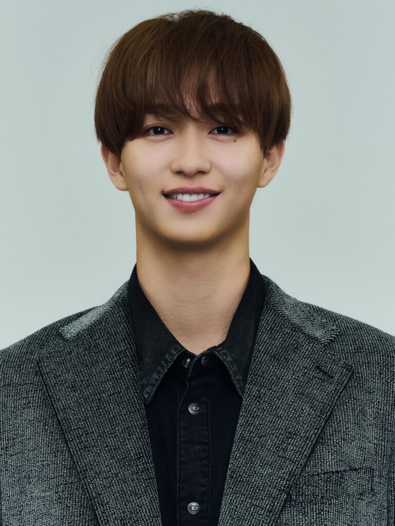 Portrait of a smiling young man with short brown hair, wearing a gray tweed blazer and black shirt against a pale background.