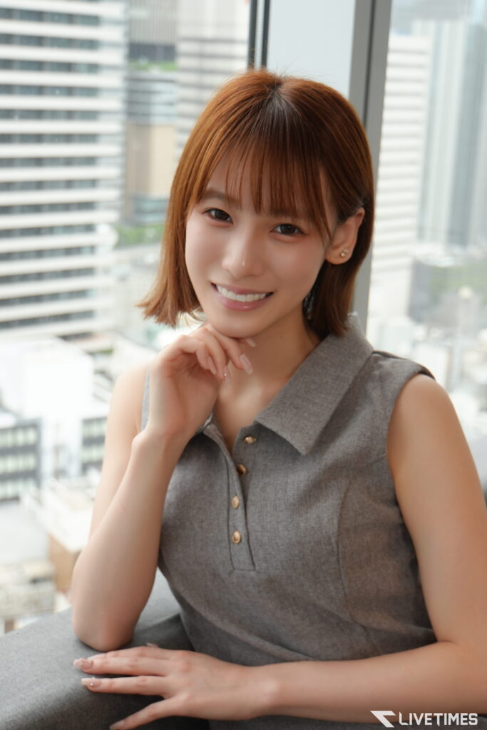 Smiling woman with short brown hair leaning on a windowsill, city buildings visible outside the window.