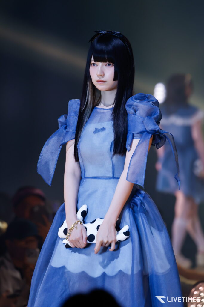 Female model in a blue satin dress with puff sleeves, holding a white polka dot clutch on a runway.