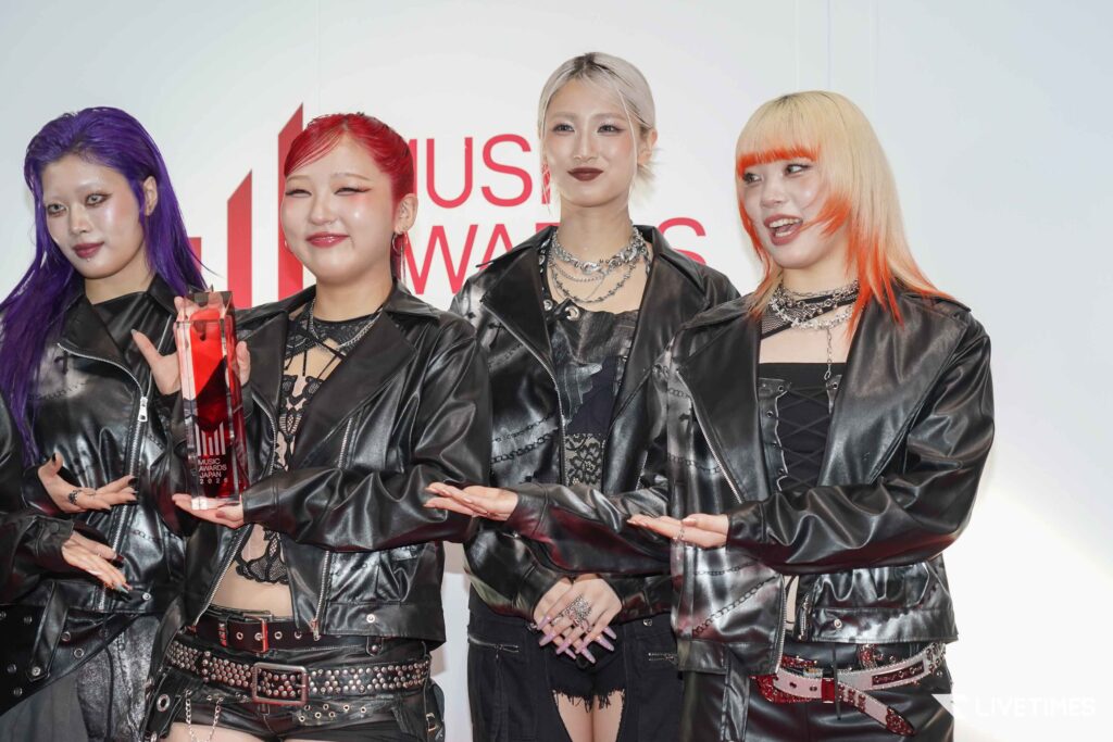 Four women in black leather outfits posing together on stage, holding a tall red crystal award at the Music Awards Japan 2026 attendance backdrop behind them.