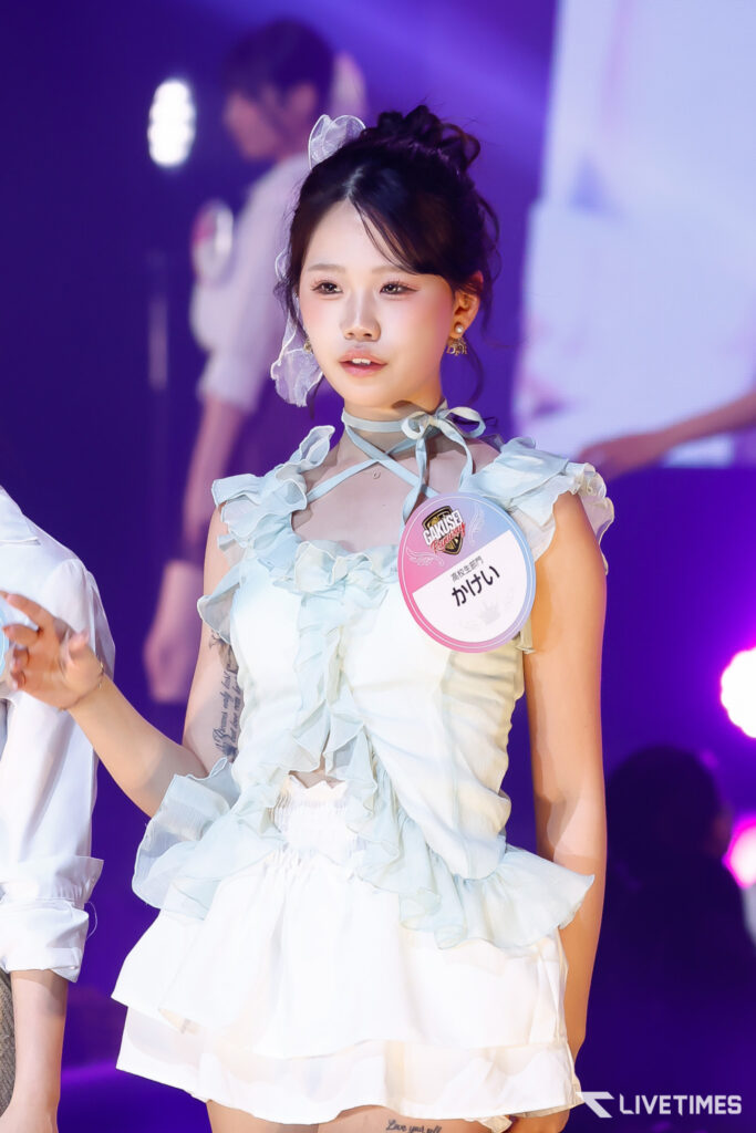 Young woman in a pale blue ruffled dress on stage with a round badge, purple lights in the background.