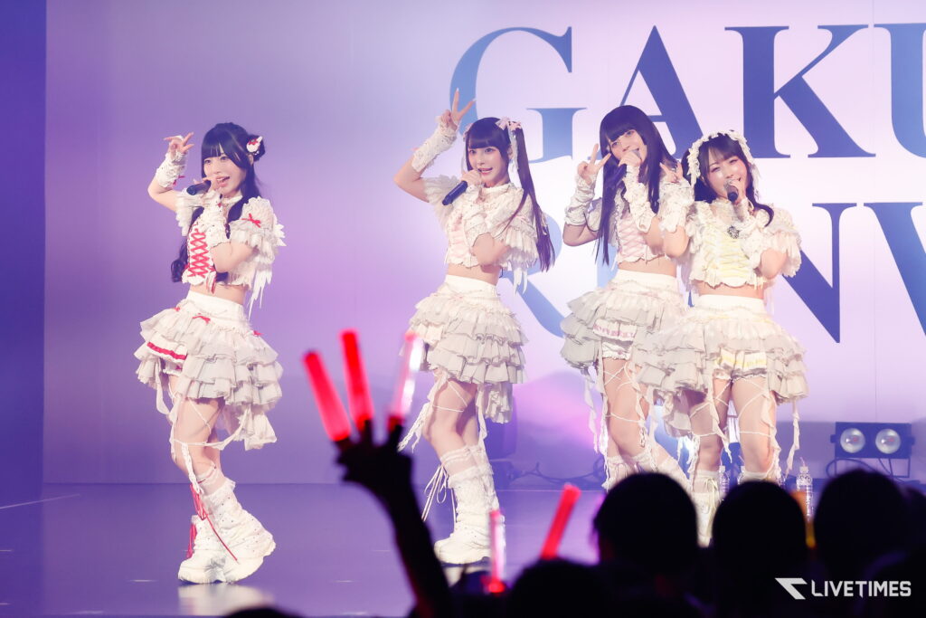 Four female performers in white frilly outfits sing into microphones on stage, with audience members waving red glow sticks in the foreground.