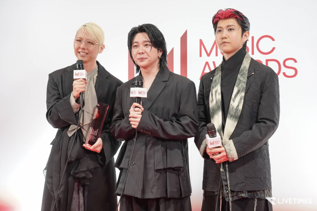 Three people in black outfits on a red-carpet stage, each holding a microphone with 'Music Awards' branding visible.