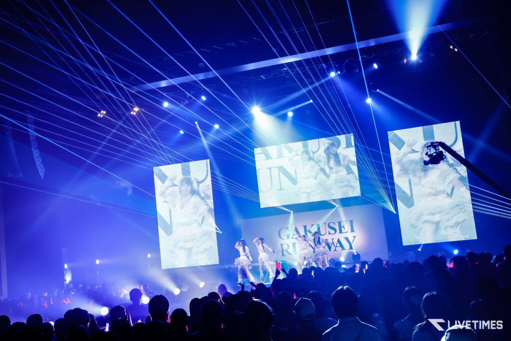 Crowd watching a blue-lit concert with laser beams and large screens showing performers on stage.