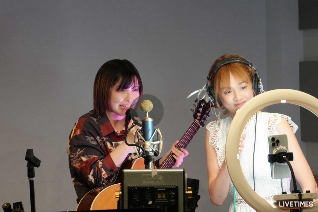 Two women recording music in a studio: one singer at a microphone with a guitar, the other wearing headphones nearby with a ring light in view.