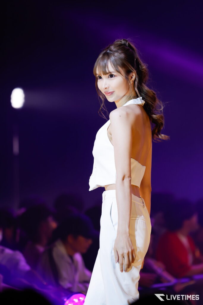 Woman in a white sleeveless outfit on stage, facing left, under purple spotlight with a blurred audience in the background.