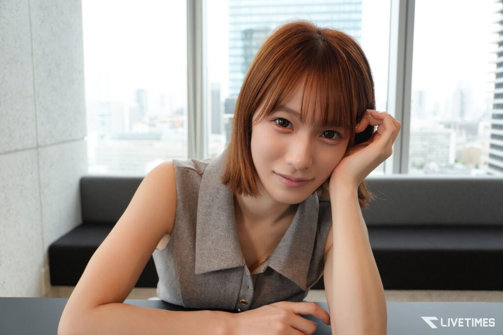 Woman with light brown hair sits at a table in a modern office, city skyline visible through windows behind her.