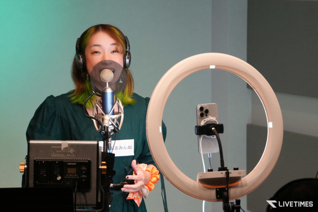 Woman wearing headphones speaks into a studio microphone with a pop filter in a recording studio environment in front of a green wall.