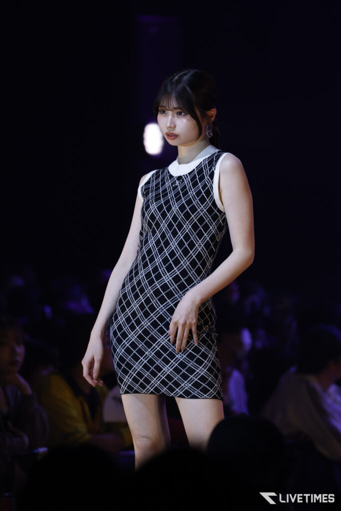 Model walks the runway in a sleeveless black-and-white plaid dress against a dark stage, audience silhouettes in the background.