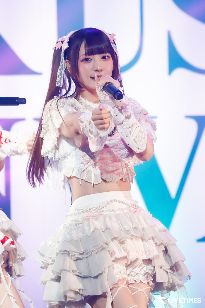 Young female singer in a white lace frilly outfit performs on stage, holding a microphone and pointing toward the audience.