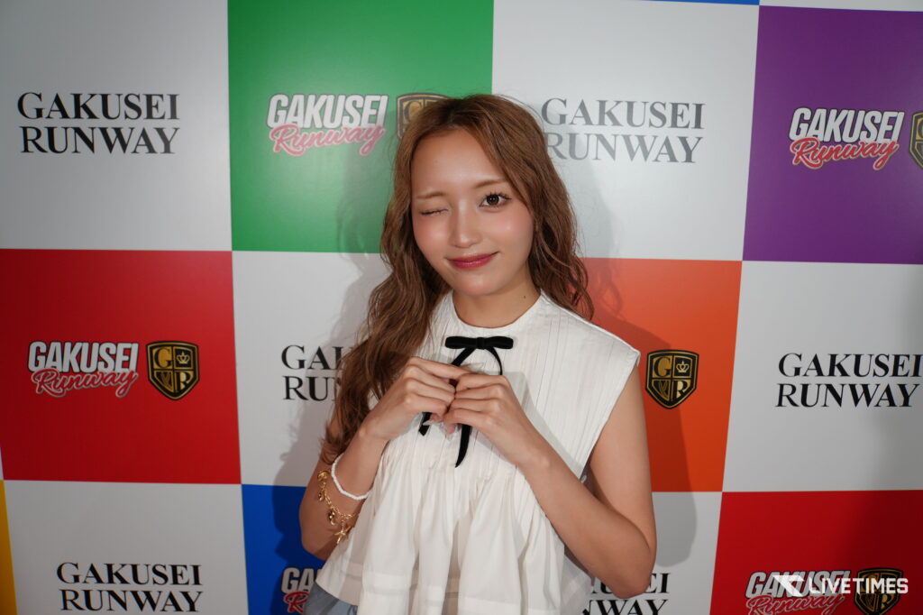 Woman posing in a white dress with black bow, winking at camera against a colorful ‘Gakusei Runway’ backdrop.