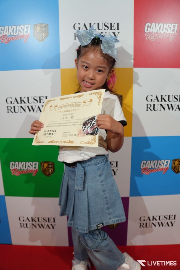 Young girl posing with a certificate at a GAKUSEI RUNWAY event, wearing a blue bow and denim skirt in front of a colorful backdrop.