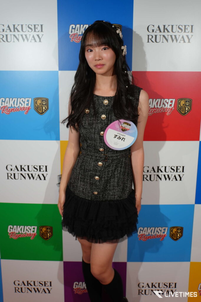 Young woman posing at a Gakusei Runway event, wearing a black textured dress with gold buttons and a round badge.