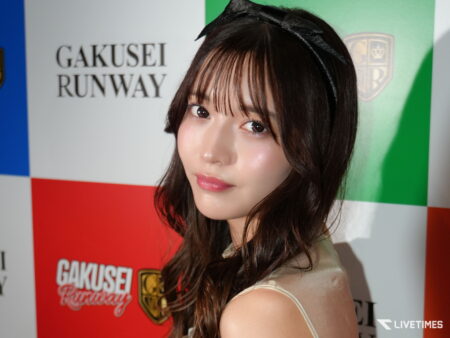 Close-up portrait of a young woman with long dark hair and glossy makeup posing in front of a colorful backdrop with 'GAKUSEI RUNWAY' text nearby.