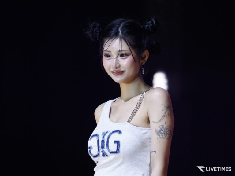 Portrait of a female performer on stage, wearing a light gray tank top, chain strap, and hoop earrings, with tattoos on her arm against a dark background.