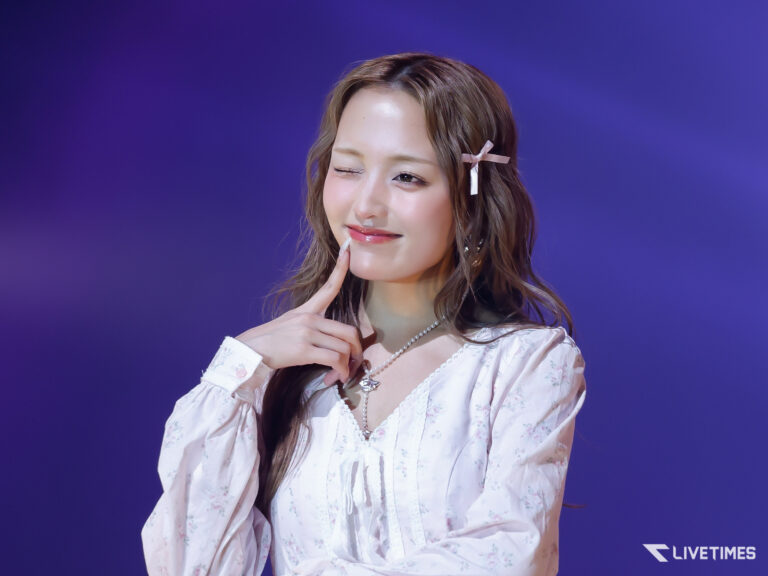 Woman winking and touching her chin, wearing a floral white blouse and pearl necklace against a purple background.