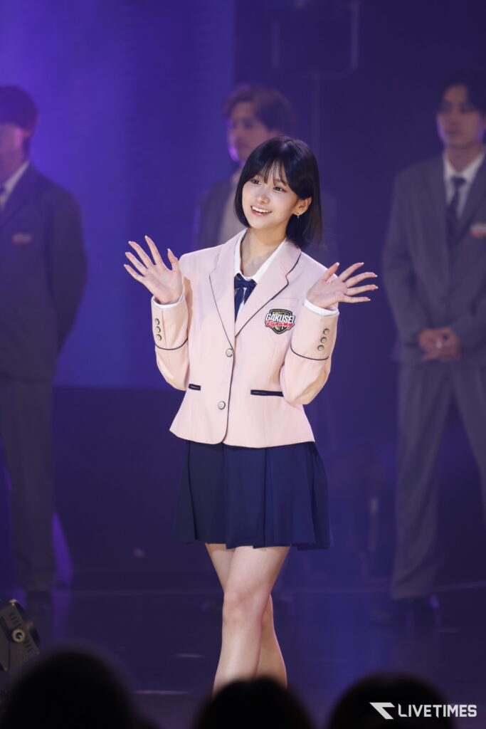 Young woman in a pale pink blazer and navy skirt raises hands in a stage performance with purple lighting in the background.